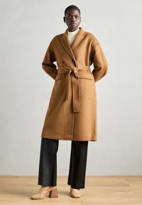 Brauner Wollmantel mit Taillengürtel, Reverskragen und zwei Taschen vorne, getragen über einem cremefarbenen Pullover und schwarzen weiten Hosen. Strukturierte, hochhackige Stiefel.