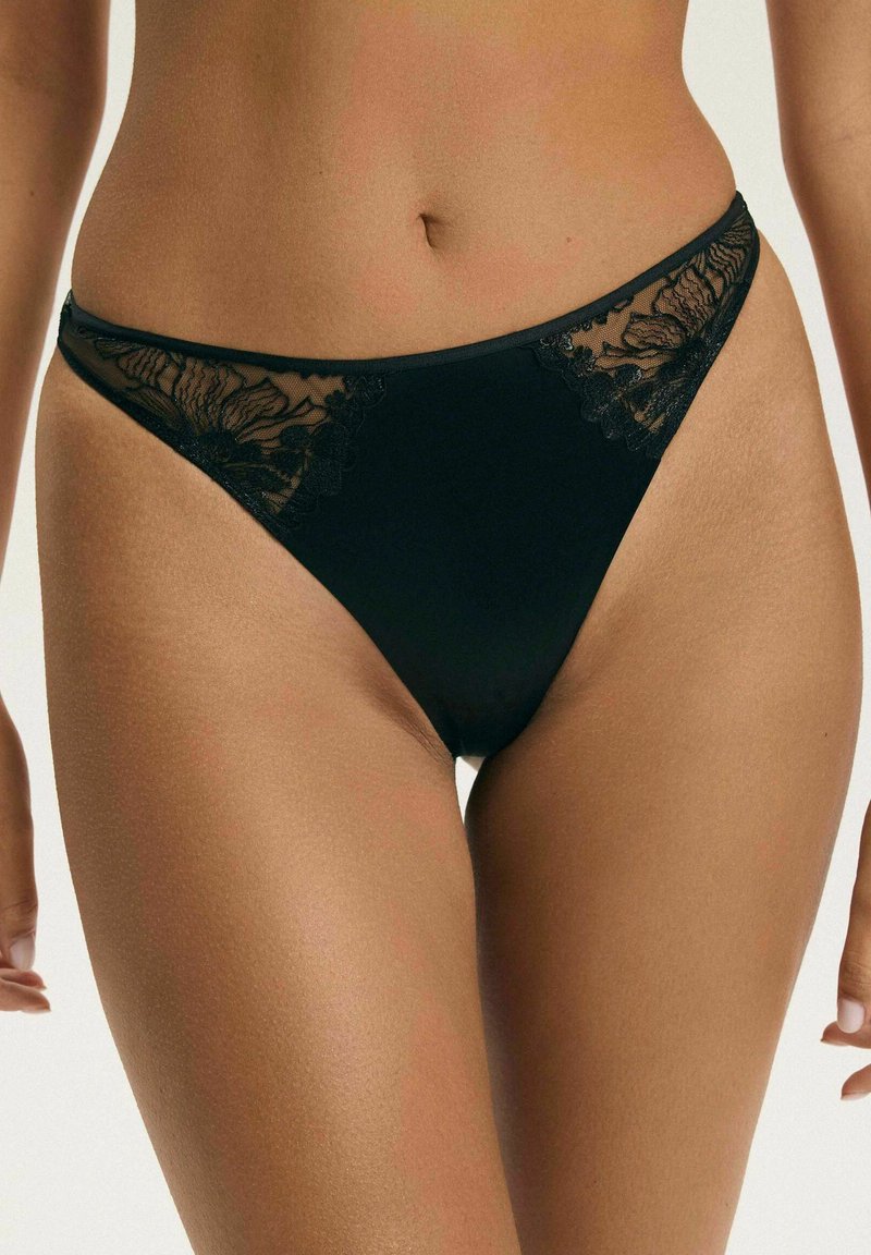 Czarne majtki z koronkowym wykończeniem noszone na biodrach kobiety, odsłaniające część jej torsu i górne uda na neutralnym tle.