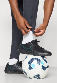 Fekete sportcipők texturált felülettel, türkiz kiegészítőkkel és fekete sportnadrággal. Egy fehér focilabda kék és fekete mintákkal a talajon.