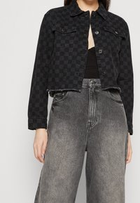 Woman wearing a cropped black checkered denim jacket over a black top with high-waisted faded gray wide-leg jeans.