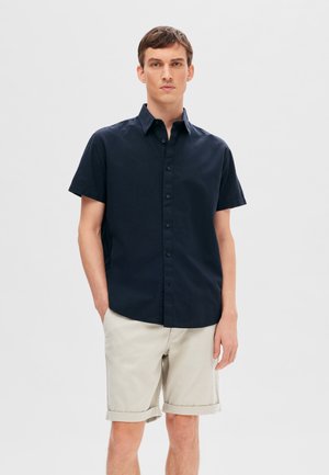 Camicetta a maniche corte blu navy con colletto button-down, realizzata in tessuto intrecciato. Abbinata a pantaloncini chiari beige arrotolati. Design semplice e strutturato.