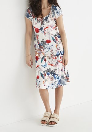 Frau trägt knie- bis knöchellanges weißes Kleid mit bunten Blumen- und Schmetterlingsmustern, steht vor einer weißen Wand, beige gestrickte Sandalen.