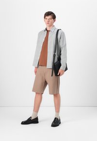 Grå rutete skjorte med knapper over en brun t-skjorte, sammen med beige ribbede shorts og svarte slip-on sko. Svart skulderveske.