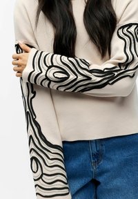 Beiger Pullover mit schwarzen abstrakten Mustern entlang der Ärmel, mit lockerer Passform und weicher Textur. Getragen mit blauen Jeans aus Denim.