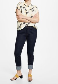 Chemise jaune à manches courtes avec motif d'ananas noir et floral, associée à un jean bleu foncé ajusté et des sandales à talons jaunes.