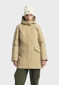 Parka impermeabile beige con cappuccio, coulisse, due tasche frontali e taglio dritto. Indossata sopra pantaloni cargo verdi e un berretto color crema.