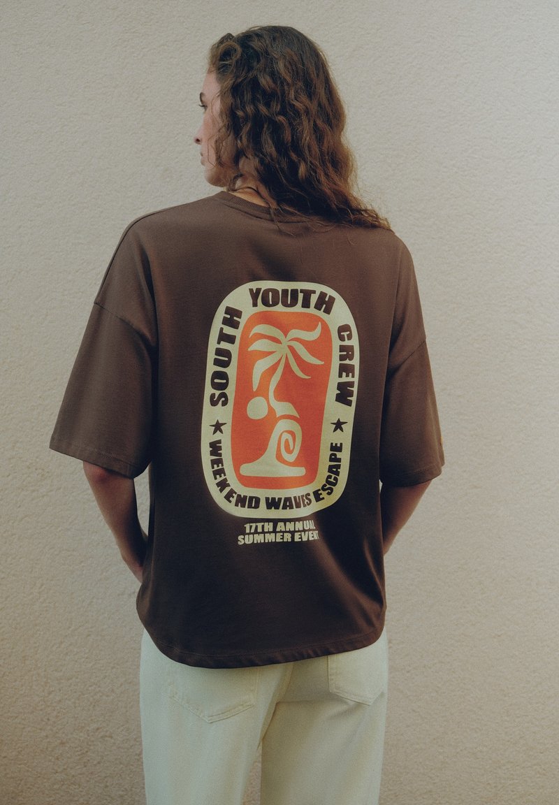 Personne aux longs cheveux bouclés tournée de côté, portant un t-shirt marron avec un graphisme coloré et un texte au dos.