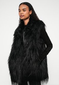 Colete de pelúcia sintética preto com uma textura longa e desgrenhada, apresentando um fecho frontal com zíper e colarinho alto. Usado sobre uma blusa preta de manga longa.