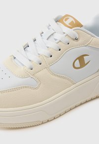 Baskets blanches et beiges avec un dessus en daim texturé, des accents en cuir lisse, une semelle en caoutchouc épaisse et un logo doré sur la languette.