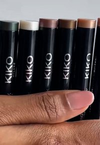 Set van KIKO Milano make-up stiften, met verschillende aardetinten in draaibare tubes met zwarte verpakking en zilveren opdruk. Hand houdt ze vast.