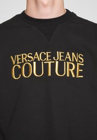 Svart sweatshirt med fet guld broderad text "VERSACE JEANS COUTURE," rund hals och texturerad tyg.