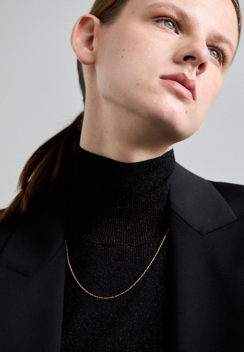 Pulover negru cu guler înalt, asortat cu un sacou negru, având un lănțișor delicat din aur; textură fină și design elegant.