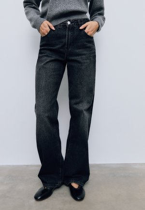 Persoon draagt zwarte wijde broekspijpen met hoge taille, handen in de zakken, grijze trui en zwarte platte schoenen, staand tegen een witte achtergrond.