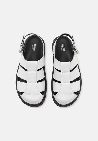White leather sandals with a strappy design, black accents, adjustable buckle, and a textured sole. Inner branding visible on the footbed.