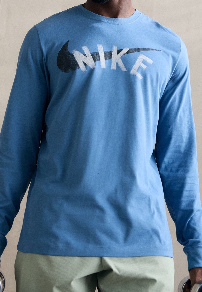 Muž oblečený v modré košili s dlouhým rukávem značky Nike a světlých kalhotách, držící činky po svých stranách na jednobarevném pozadí.