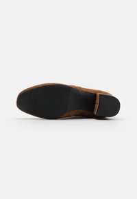 Suela de zapato de ante marrón con agarre de goma negra texturizada y diseño de punta cuadrada sobre un fondo blanco.