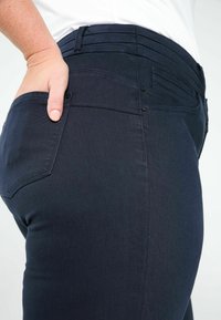 Jean en denim bleu foncé avec une texture lisse, coupe ajustée et style à cinq poches. Comprend une fermeture par bouton et des accents de couture visibles.