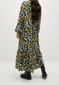 Pitkä, kukkakuvioinen mekko mustalla taustalla, jossa on keltaisia, valkoisia ja vihreitä kuvioita. Mekossa on puffihihat, kerroksellinen helma ja se on yhdistetty mustiin saappaisiin.