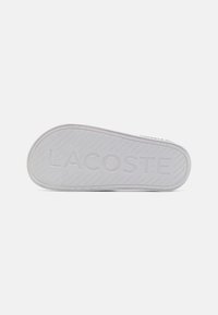 Semelle en caoutchouc blanche avec le texte "LACOSTE" embossé et un motif de semelle diagonale pour l'adhérence ; texture lisse avec des bords arrondis.