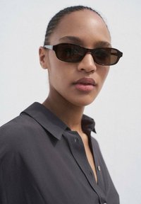 Occhiali da sole con montatura rettangolare in marrone scuro, caratterizzati da asticelle sottili e una finitura lucida. Indossati con una camicia button-up scura.