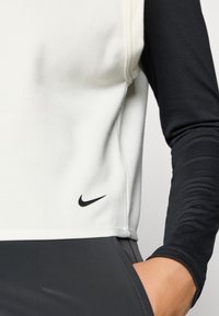 Nike sporditopp, mis on valge ilma varrukateta disainiga ja mustade pikkade varrukatega. Pehme tekstuur, külje logo detail ja õmblusteta konstruktsioon.