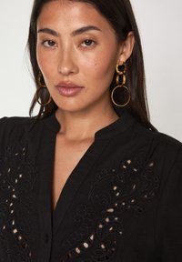 Chemise noire avec un col mandarin, ornée de broderies complexes et de découpes. De grandes boucles d'oreilles en or complètent l'ensemble.