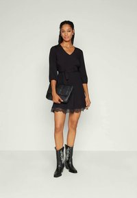 Μαύρο φόρεμα με δαντελωτή διακόσμηση, V-λαιμόκοψη και ζωνάκι στη μέση. Το μοντέλο φοράει μαύρες μπότες μέχρι το γόνατο με άσπρα λουλουδάτα μοτίβα και κρατάει μια μαύρη τσάντα.