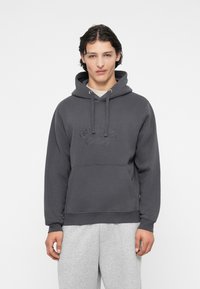 Sudadera gris con bolsillo canguro, capucha ajustable con cordón, y logo en relieve. Tejido suave con un corte relajado y puños de canalé.