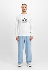 Langermet hvit T-skjorte med svart "Alpha Industries"-logotyp, kombinert med lyseblå løse jeans og sølvfargede sneakers.