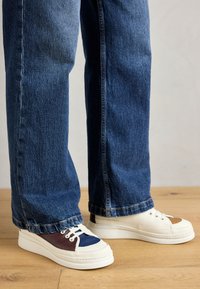 Hvide sneakers med marineblå og brune detaljer, rund tå, snørebånd foran og tyk hvid sål. Båret med blå denim jeans. Trægulv baggrund.