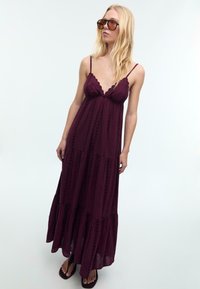 Robe maxi bordeaux avec des fines bretelles, encolure évasée, design à plusieurs niveaux et détails brodés. Associée avec des tongs marron et des lunettes de soleil.