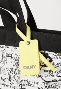 Čierna pletená taška s bielymi kreslenými vzormi, obsahujúca žltú menovku na batožinu s nápisom "DKNY" v striebornom písme. Pevné držadlá a textúrovaný povrch.