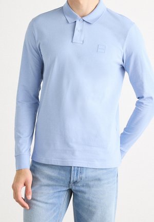 Mann trägt ein hellblaues langärmeliges Poloshirt und hellblaue Jeans, steht mit einer Hand hinter dem Rücken.
