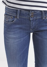 Mörkblå denimjeans med en lätt blekning; tvåknappsstängning; synlig sömnad; framfickor; "Pepe Jeans"-logotyp på vänster ficka.