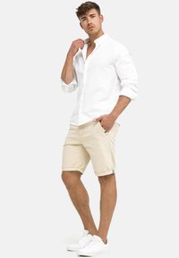 Witte button-down shirt van linnen stof, beige opgerolde shorts en witte sneakers. Schone lijnen, casual pasvorm en minimale ontwerpelementen.
