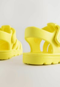 Bright yellow sandals with cut-out design, made from flexible material. Features adjustable straps and a textured sole for grip.