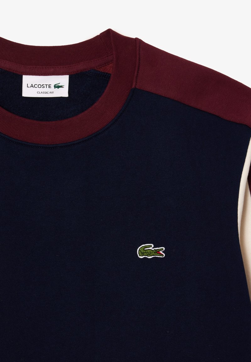 Sudadera navy con acentos burdeos y crema. Cuello de canalé, con un pequeño logo de cocodrilo bordado en verde en el pecho. Corte clásico.