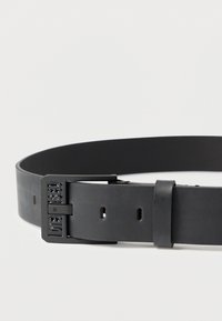 Čierny kožený opasok s matným povrchom, vybavený obdĺžnikovou kovovou prackou s vyrazeným názvom značky a viacerými dierkami na nastavenie veľkosti.