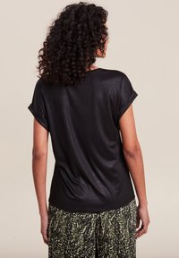 Top nero a maniche corte con risvolti, caratterizzato da una texture liscia e leggermente lucida. Abbinato a pantaloni con motivo fogliato, visto da dietro.