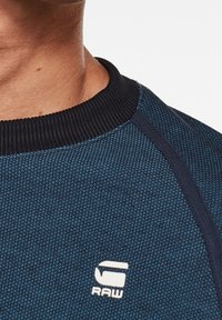 Tmavě modrá texturovaná mikina s žebrovaným kulatým výstřihem a kontrastními černými detaily s bílým logem "RAW" na hrudi.