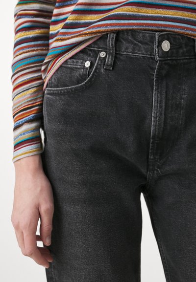 Jeans de mezclilla negra con un ajuste slim, que presentan un diseño clásico de cinco bolsillos y cierre de botón metálico. Camisa de manga larga a rayas multicolor visible.