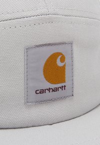 White fabric cap featuring a gray rectangular patch with a yellow logo and "carhartt" in maroon text, highlighting its textured surface.
