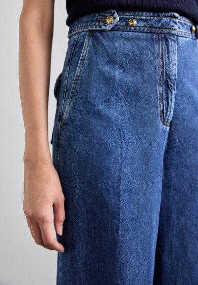 Blå denim vid benbukser med høj talje, med guld knapdetaljer og en glat tekstur. Frontlommer er synlige.
