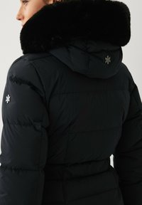 Zwarte pufferjas met een nep-bont gevoerde capuchon, voorzien van een gestikte uitstraling. Inclusief een klein embleem op de schouder en een toegesnoerde taille.