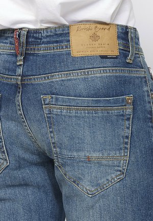 Vaqueros de mezclilla en lavado azul medio; las características incluyen un parche de cuero en la cintura, costuras dobles y dos bolsillos traseros con remaches.