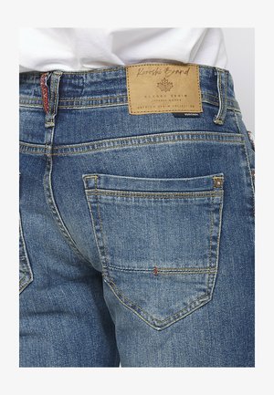 Közepesen kék mosású farmernadrág; jellemzői közé tartozik a bőr folt a derékrészen, a dupla varrás és két hátsó zseb, amelyeket szegecsek díszítenek.