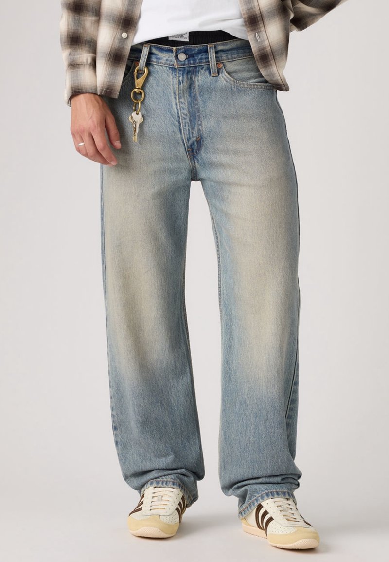 Homme portant un jean bleu délavé à jambes larges, des baskets blanches et une chemise à carreaux, avec des clés attachées à un passant de ceinture.