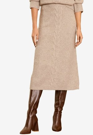 Eine Frau präsentiert einen beigen Strickpullover und einen passenden Midi-Rock, kombiniert mit stilvollen braunen kniehohen Stiefeln, und steht selbstbewusst.