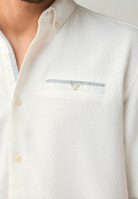 Chemise blanche à manches longues avec un motif texturé, dotée d'une poche poitrine avec un liseré bleu et un accent de bouton décoratif.