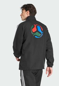 Sort jakke med høj krave. Har et stort, flerfarvet cirkulært logo på bagsiden. Inkluderer hvide striber på ærmerne.